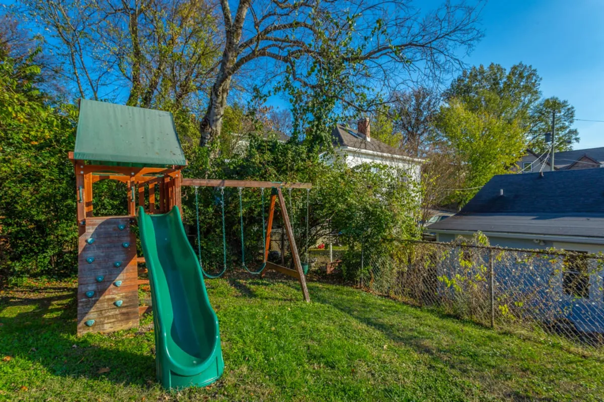 A vibrant scene featuring playground equipment including a green slide and swing set, surrounded by lush trees. Children are playing and families are gathered in the background, creating a joyful and energetic atmosphere perfect for outdoor family fun.