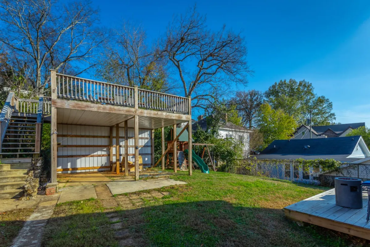 A wide exterior view highlighting a spacious deck, swing set, green grass, and patio area surrounded by trees. The space is ideal for families or groups to gather, play, or relax outdoors.