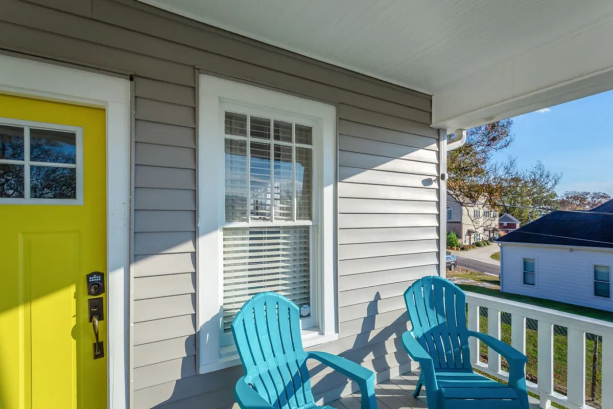 A relaxed exterior view showcases the yellow front door and blue chairs on the porch, with a window set behind. The setting suggests a peaceful spot to unwind, enjoy a coffee, or chat before heading out to explore the city.