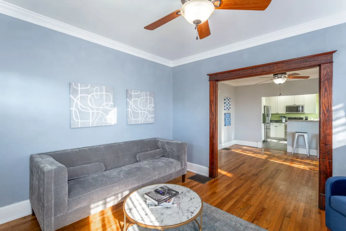 A welcoming living room with a gray velvet sofa, marble coffee table, vibrant artwork on the wall, and an archway leading to the kitchen. Natural light streams in, creating a cozy space for relaxing, socializing, or entertaining guests.