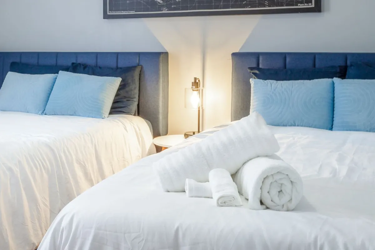 This image features two beds with blue headboards, neatly arranged towels, and a soft bedside lamp illuminating the space. The beds are made with crisp linens, and natural light filters gently into the room, creating a calm and inviting atmosphere ideal for group stays.