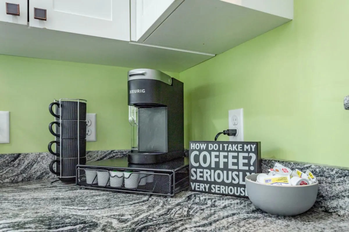 This intimate vignette shows a Keurig coffee maker, coffee pod holder, humorous coffee sign, and bowl with creamers arranged on a kitchen counter. The setup feels cozy and inviting, emphasizing the ritual of gathering for coffee.