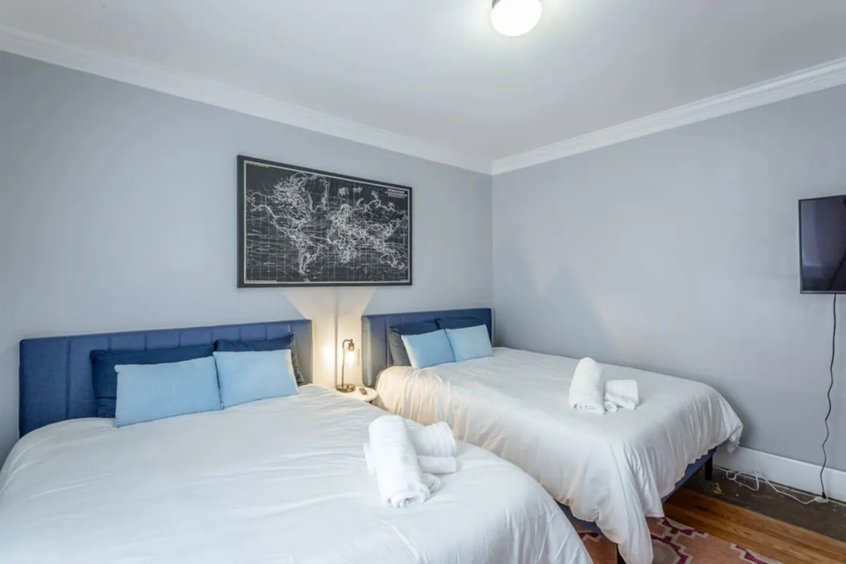 The room shows two beds with blue headboards and a world map artwork above, complemented by a wall-mounted television. The beds are neatly made, and the cozy space is designed for both rest and shared moments, offering the option to unwind with a show.