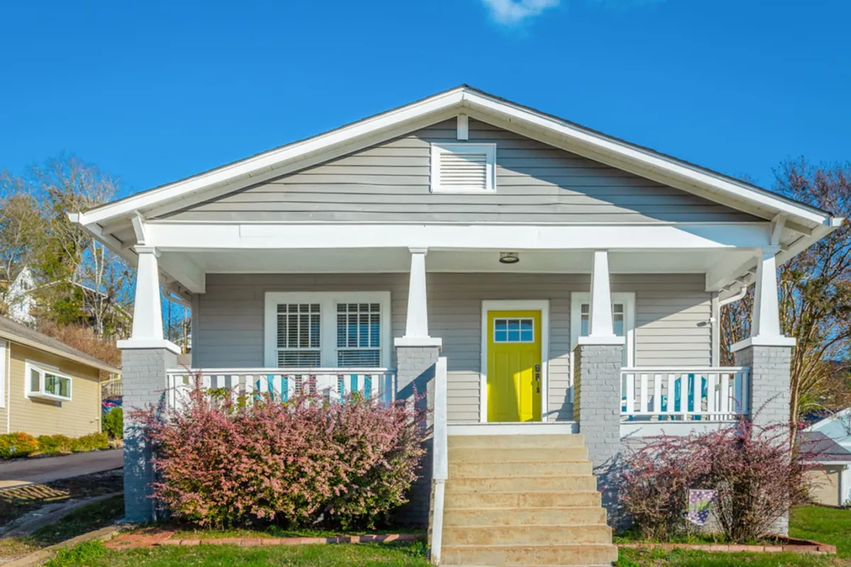 A cheerful exterior shot of the home featuring a bright yellow door, classic white porch railings, and lush landscaped shrubs. The inviting front porch is framed by greenery and bathed in natural light, creating a warm welcome for guests.