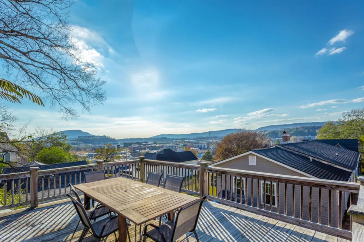 An elegant outdoor dining table sits on a spacious deck with sweeping views of the city and distant mountains, set under a clear blue sky. The setting conveys a communal, serene atmosphere ideal for group gatherings, al fresco meals, and soaking up the panorama.