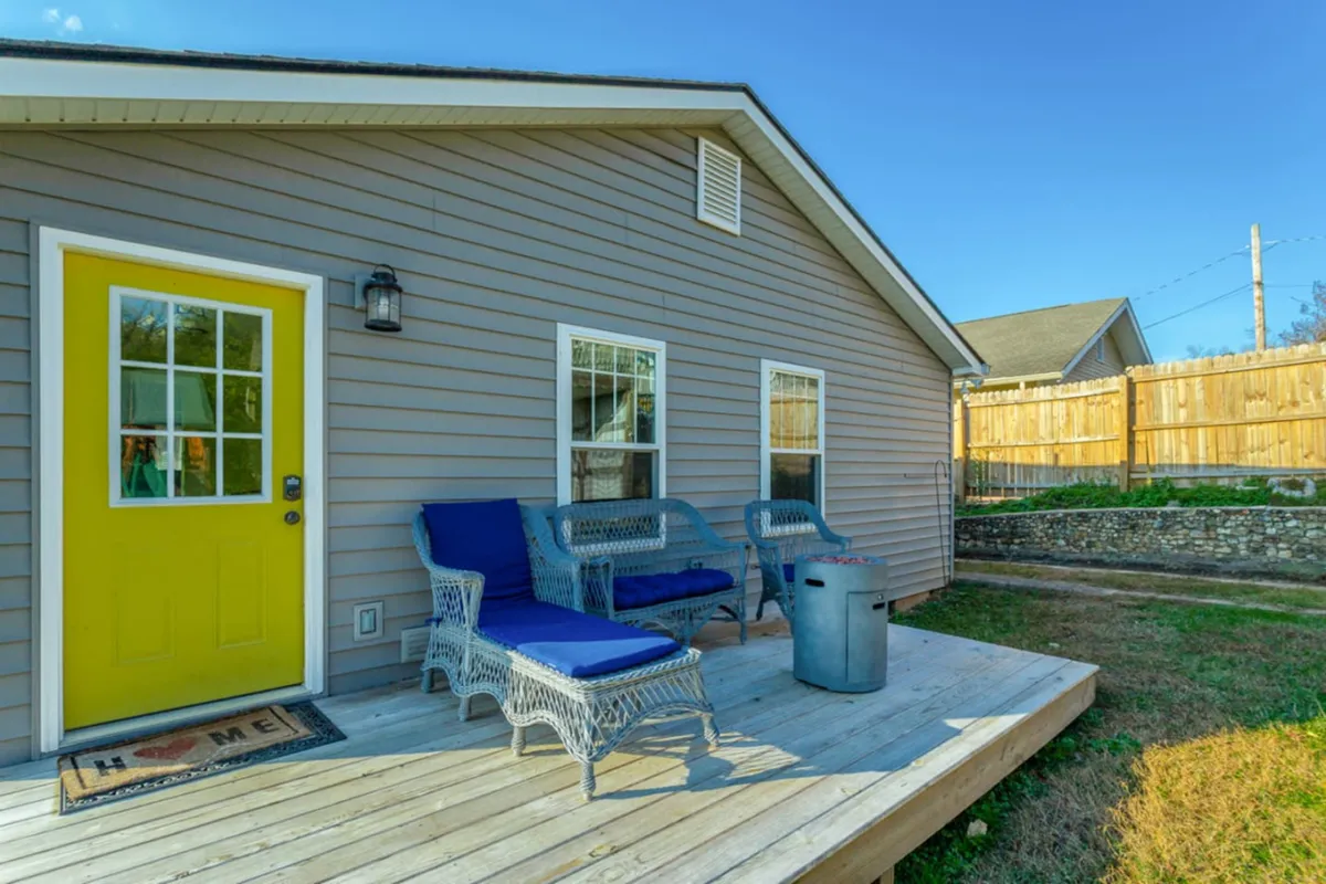 This exterior image features a welcoming yellow front door, blue chairs arranged on a wooden deck, and a glimpse of the home's inviting outdoor gathering space. The setup evokes a sense of relaxation and socializing, ideal for coffee in the morning or unwinding at sunset.