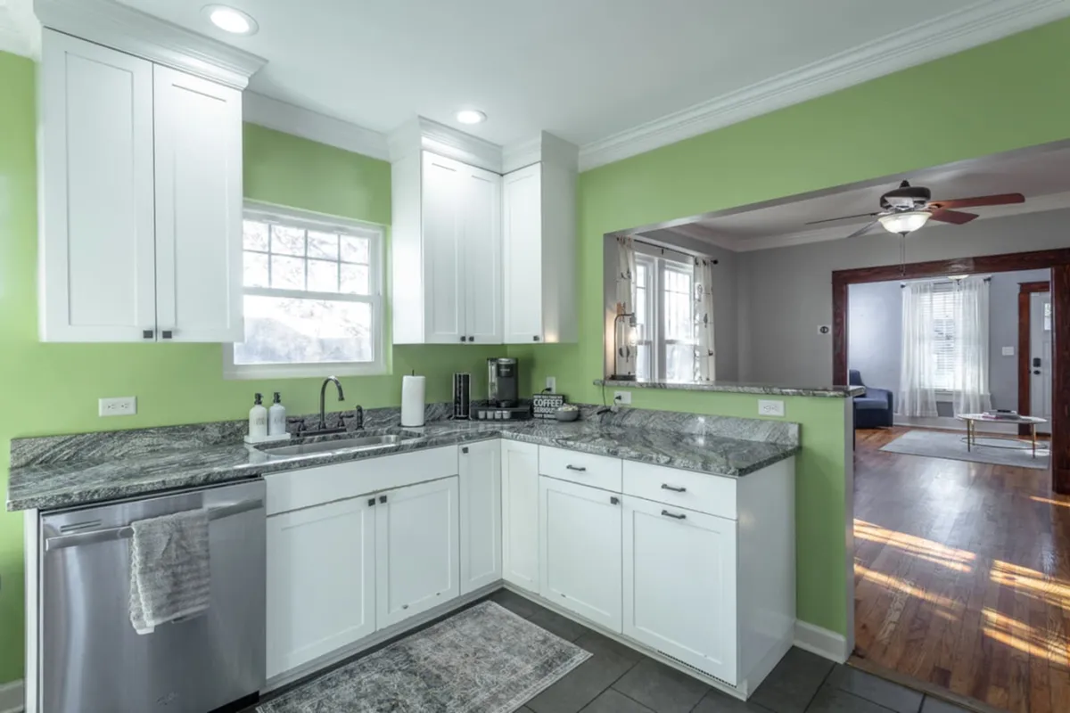 A vibrant kitchen scene with granite countertops, green walls, white cabinetry, and various kitchen appliances. Natural morning light floods the space, highlighting areas ideal for group meal prep or sharing recipes.