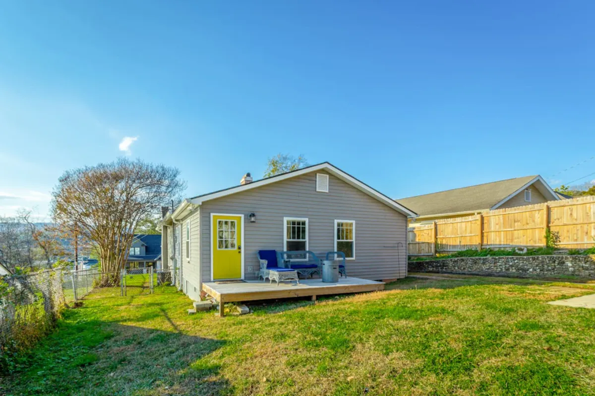 The image showcases a bright yellow door that opens onto a spacious deck area, surrounded by a green lawn and mature trees. The outdoor setting is cheerful and tranquil, with opportunities for both relaxation and social gatherings.